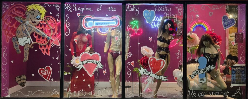 Colorful storefront window display featuring playful, romantic artwork and mannequins in lingerie, promoting inclusivity and self-expression at CRAVE Romance Boutique.