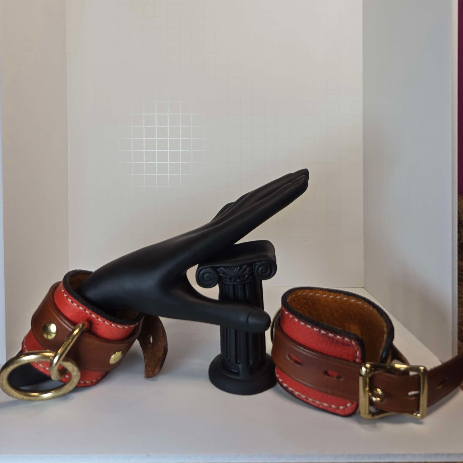 Handcrafted leather cuffs with gold hardware displayed alongside a decorative black hand and a small column, showcasing W.E.L.P.'s artisan bondage gear.
