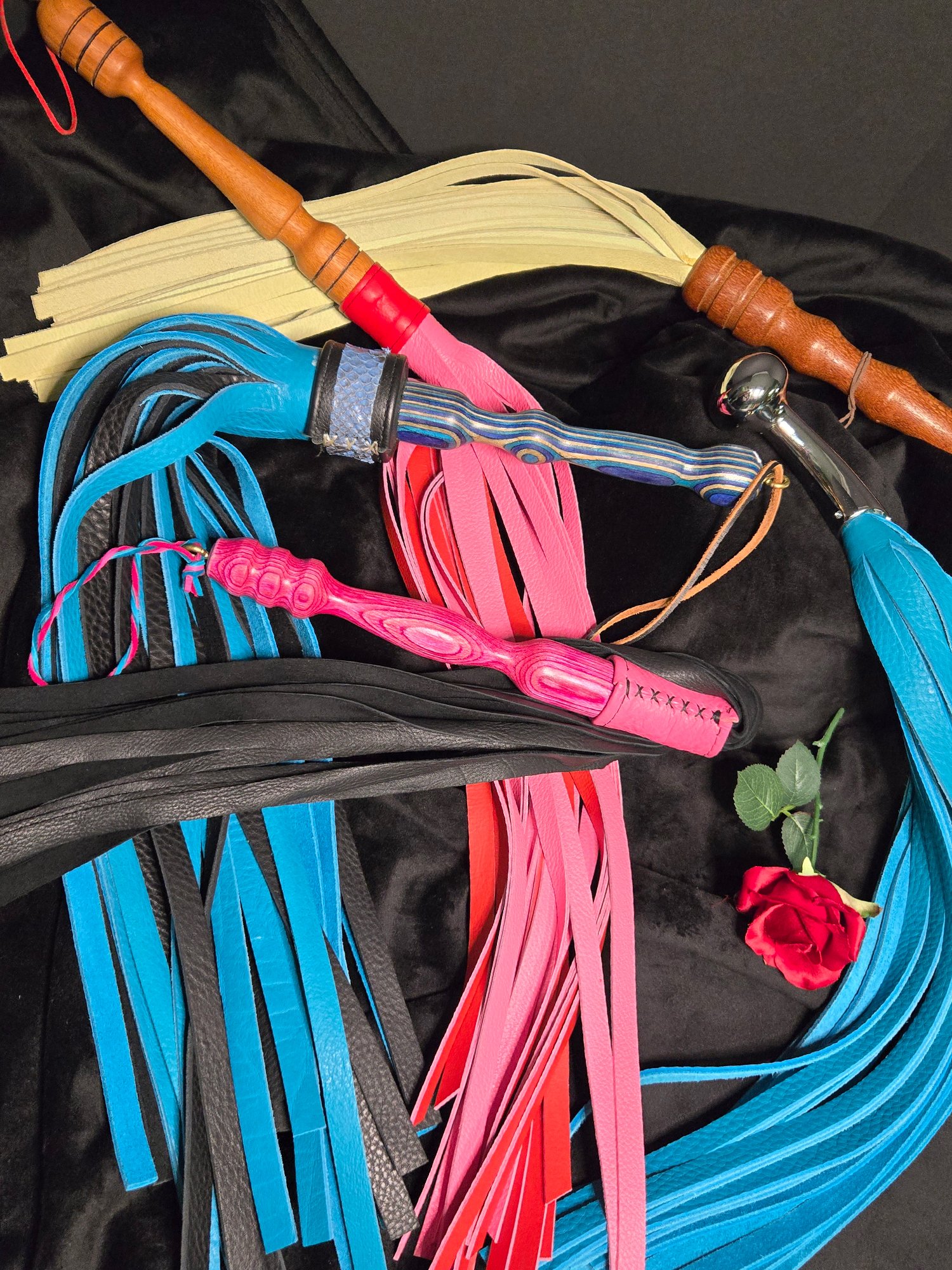 Handcrafted impact toys including custom floggers and paddles, featuring colorful leather tails and turned wooden handles, displayed on a black background with a red rose accent.