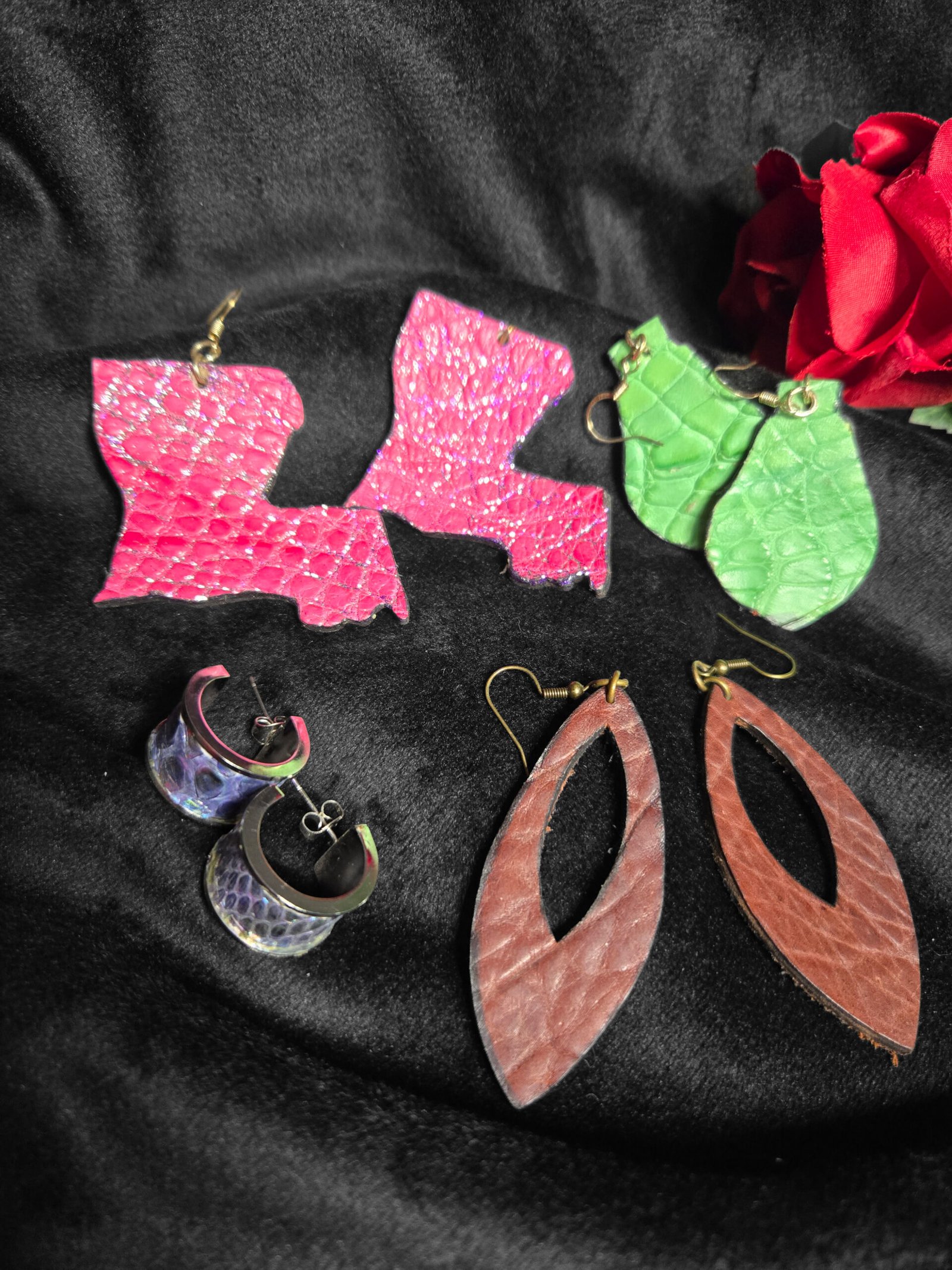 Colorful leather earrings including pink Louisiana-shaped pieces, green leaf designs, iridescent hoops, and brown oval cutouts, showcasing artisan craftsmanship from W.E.L.P.