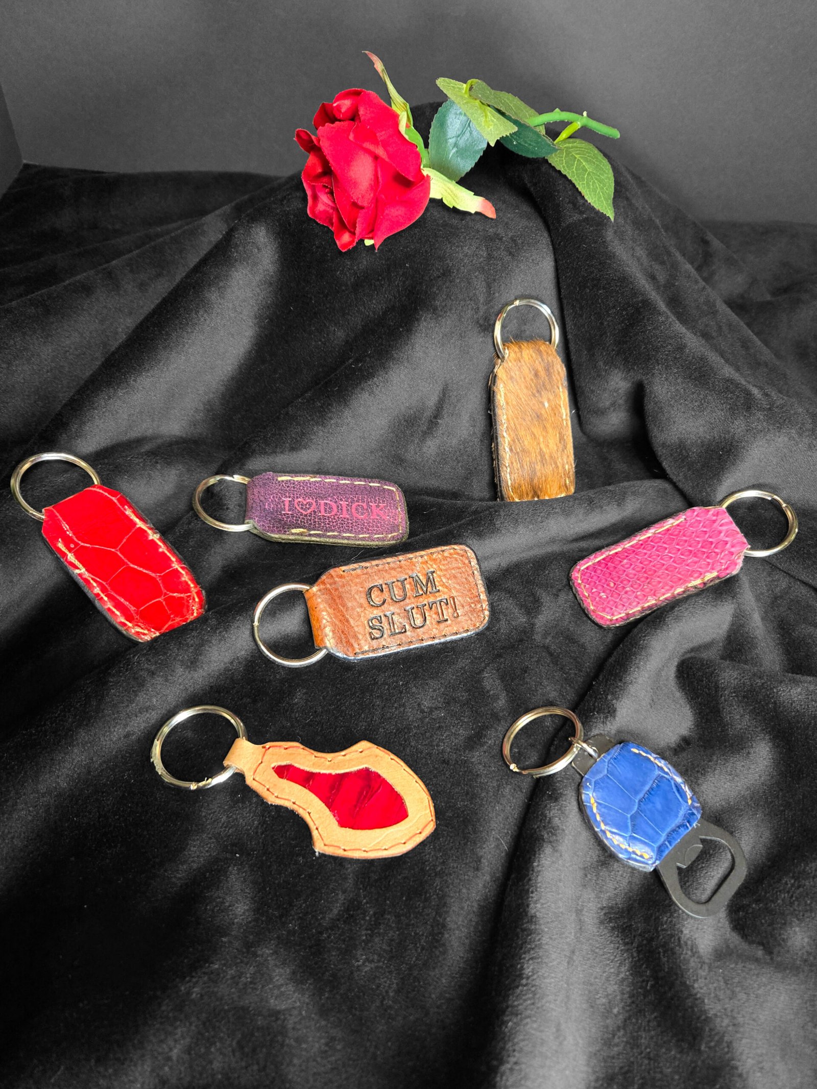 Sturdy leather keychains in various colors and designs, featuring playful phrases like "I ♥ DICK" and "CUM SLUT," alongside a red rose, on a black fabric backdrop, showcasing W.E.L.P.'s artisan craftsmanship in unique accessories.