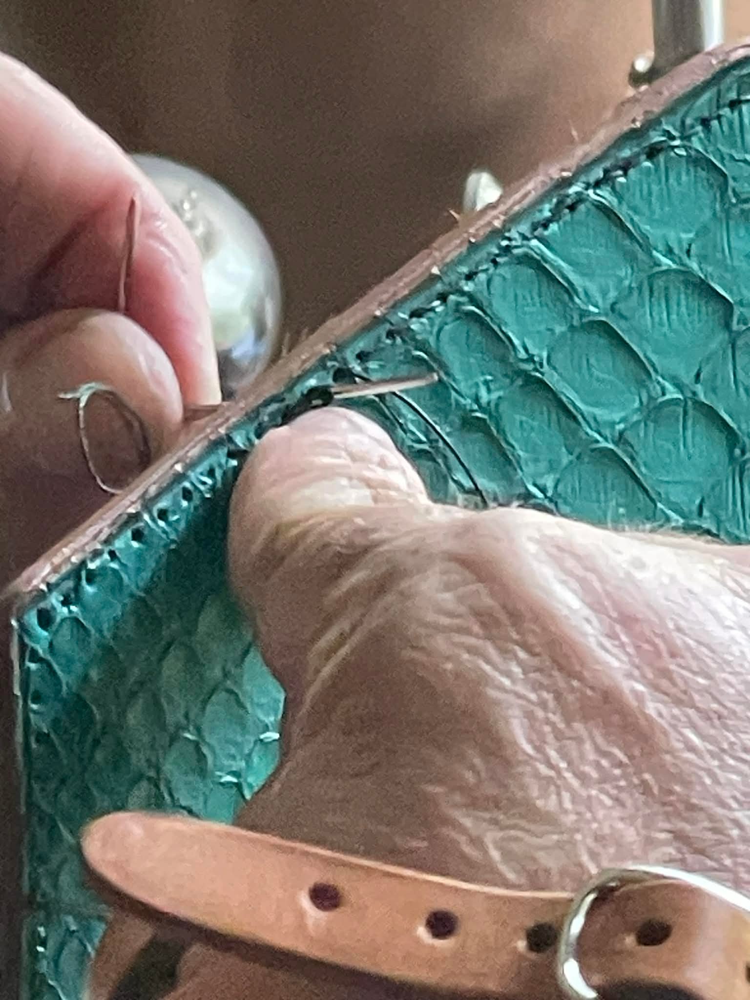 Hand holding needle and thread stitching handcrafted turquoise leather, showcasing artisanal craftsmanship in leather goods.
