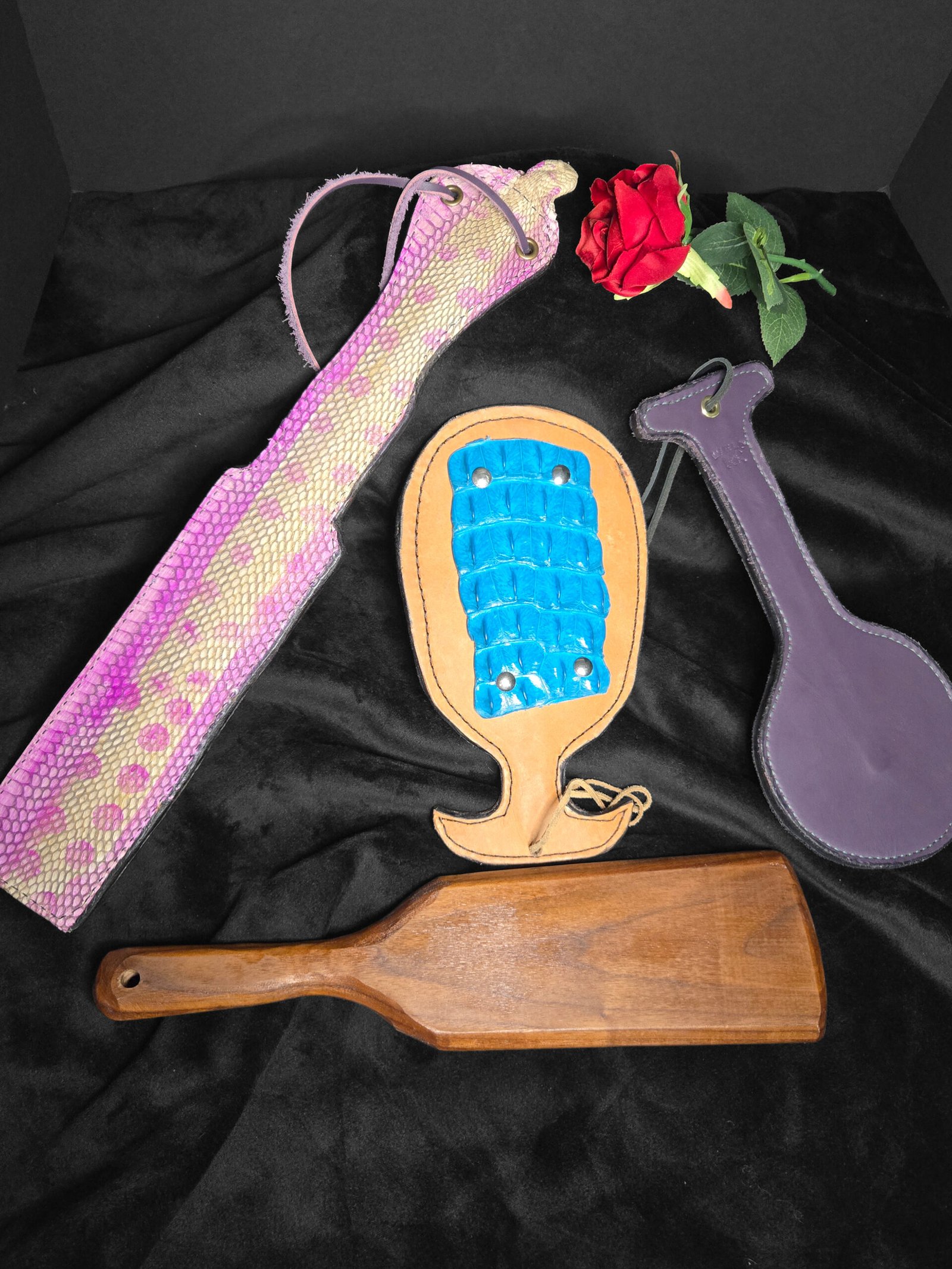 Exotic leather paddles and accessories, including a pink python paddle, a wooden paddle, and a blue-textured leather piece, arranged artistically with a red rose on black fabric backdrop, showcasing W.E.L.P.'s handcrafted BDSM gear.