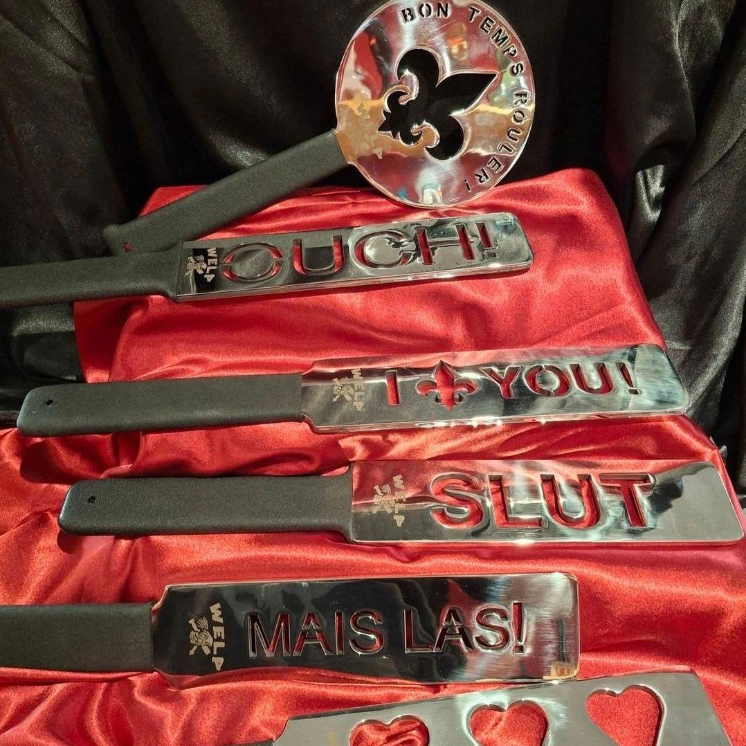 Custom metal paddles with engraved phrases, including "OUCH!", "I ♥ YOU!", "SLUT", and "MAIS LAS!", displayed on red satin fabric, showcasing W.E.L.P.'s handcrafted BDSM gear.