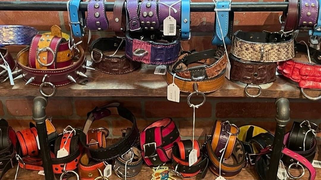 Colorful handcrafted leather cuffs and collars displayed on a wooden shelf, featuring various designs, textures, and metal embellishments, showcasing W.E.L.P.'s artisan craftsmanship in bondage wearables.