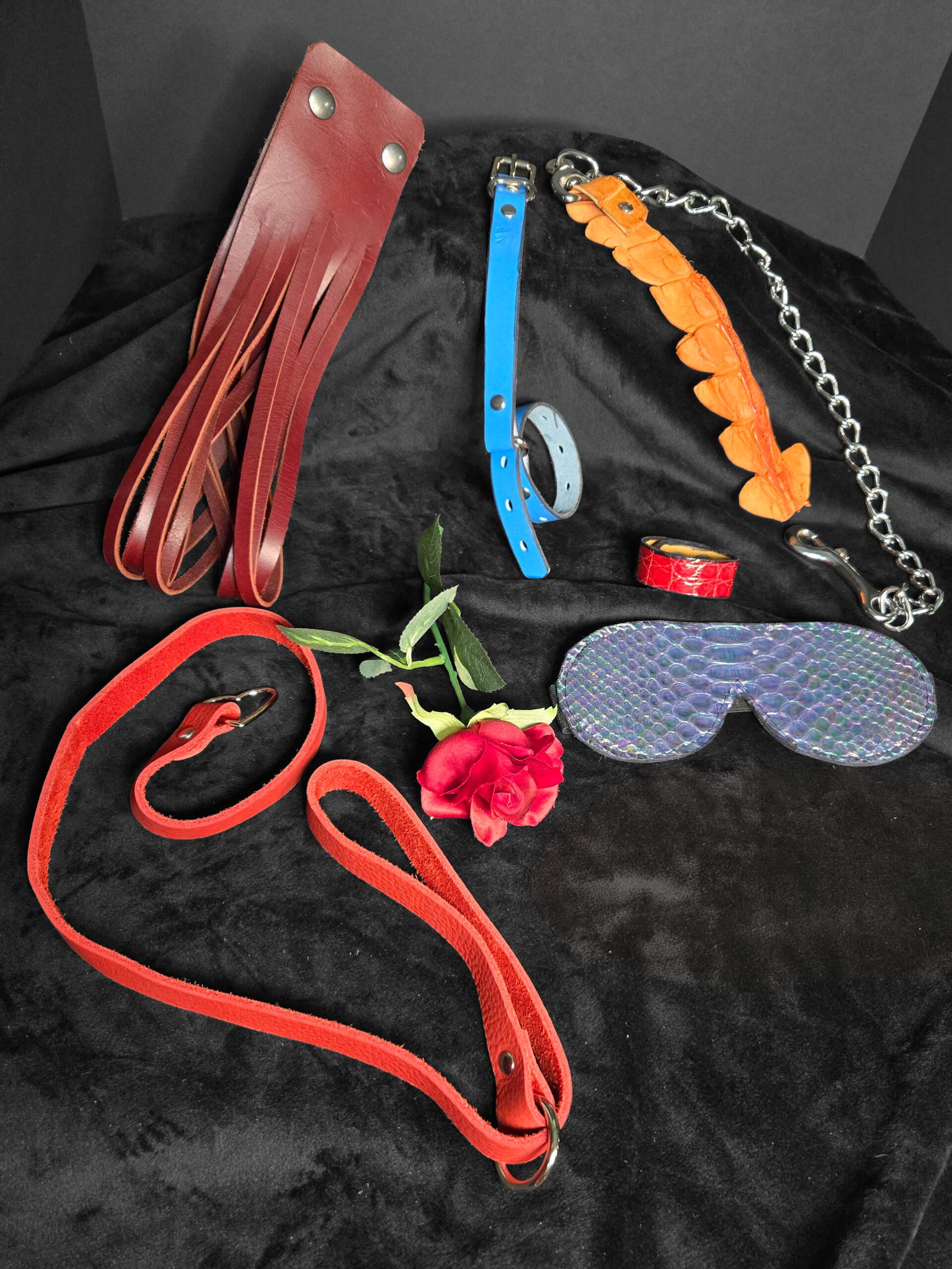 Handcrafted leather BDSM accessories including a red flogger, blue collar, orange tail whip, and patterned blindfold, arranged with a rose on a black background.