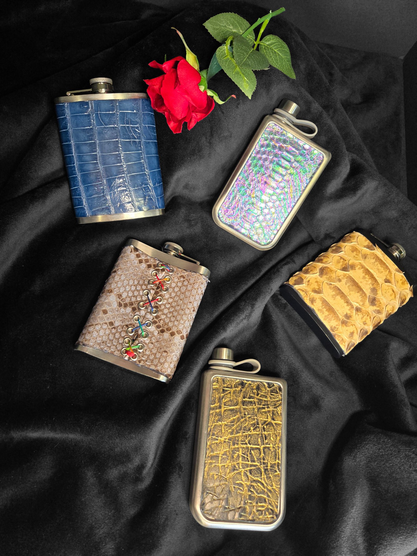 Custom-wrapped leather flasks in various textures and colors, including blue crocodile, iridescent snake, and embellished designs, arranged on a dark fabric backdrop with a red rose.