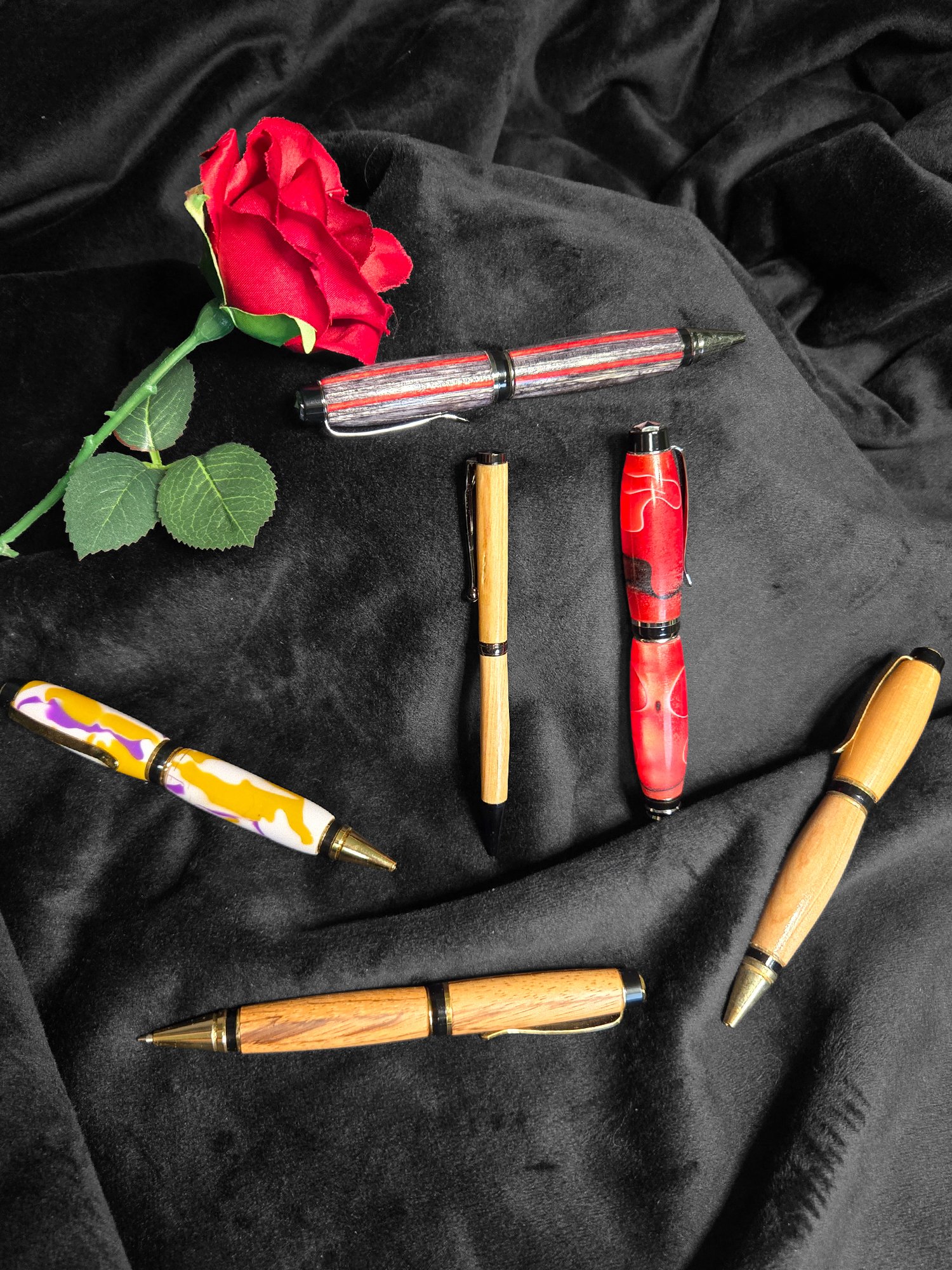 Hand-turned wood pens in various colors and designs, alongside a red rose, on a black velvet background, emphasizing artisan craftsmanship and elegant writing accessories.