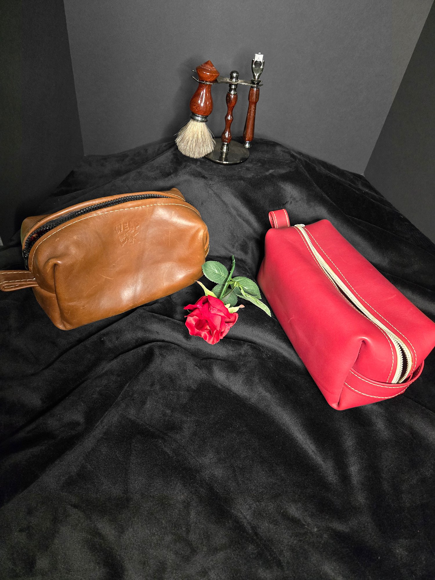 Handcrafted leather dopp kits in brown and red, elegantly displayed with a rose and traditional shaving tools on a black fabric background, showcasing W.E.L.P.'s artisan craftsmanship for travel essentials.